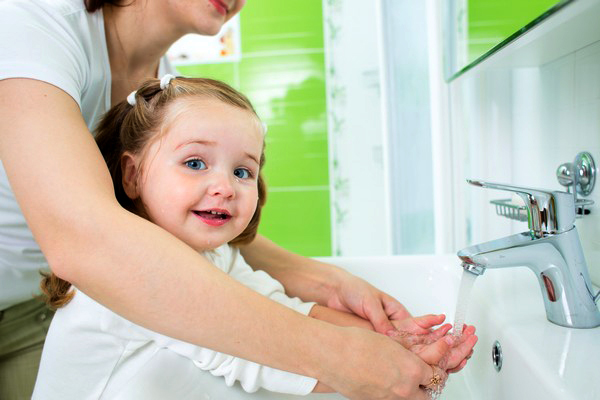 Young girl washing her hands at a sink with the help of a woman standing behind her.